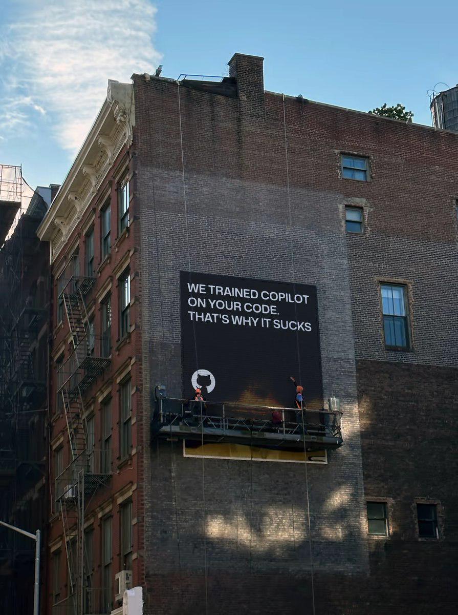 The meme is a picture of a construction worker's safety sign being removed from a high-rise building. The sign reads: "We trained a copilot on your code. That's why it sucks."